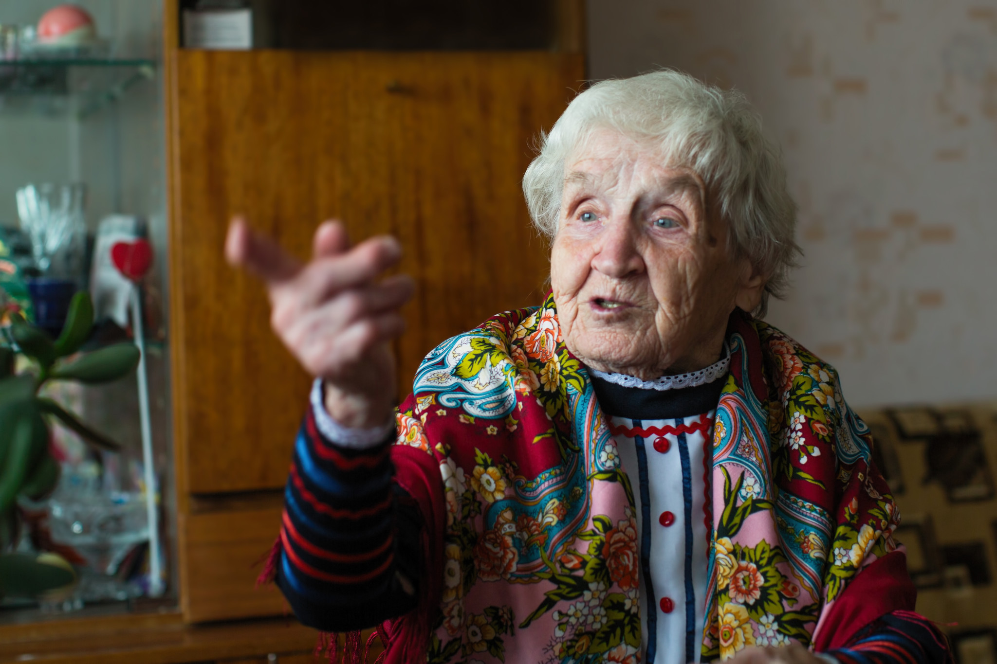An old woman in bright clothes and a scarf gestures sitting at the table. Elderly pensioner.
